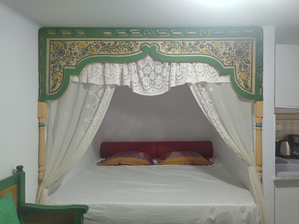 Cama con dosel decorada con detalles coloridos y cortinas de encaje en un apartamento típico frente al mar en Mahdia.