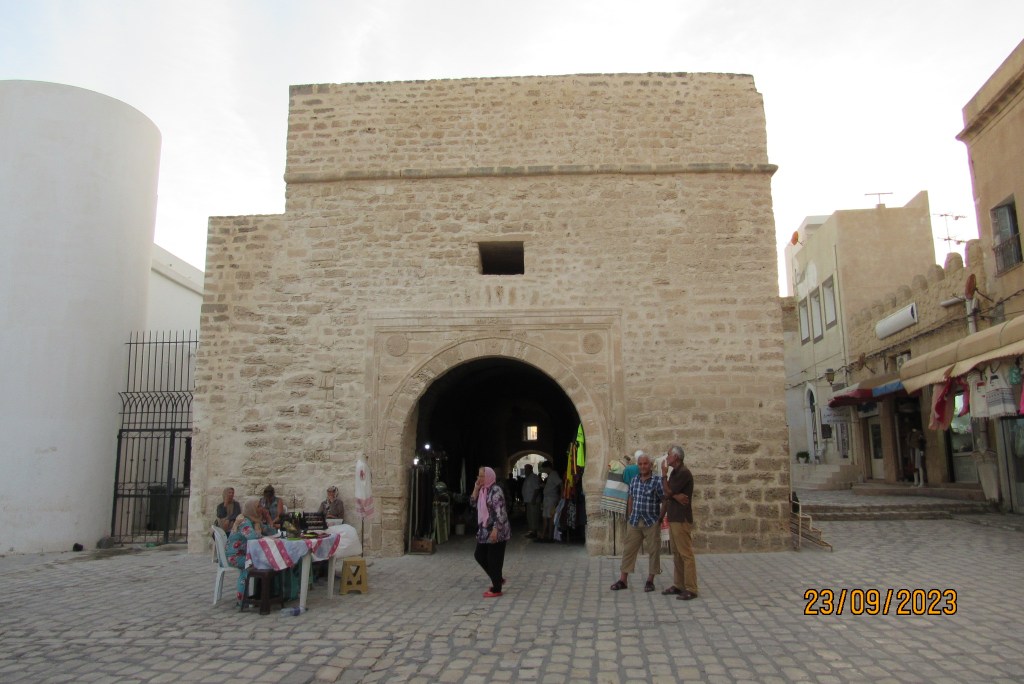 Vista de una antigua puerta de entrada en Mahdia, con personas conversando y un ambiente pintoresco.